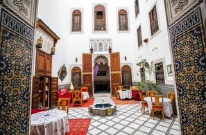 Um pátio central com uma fonte, rodeado por mesas e cadeiras, e portas e janelas decoradas.