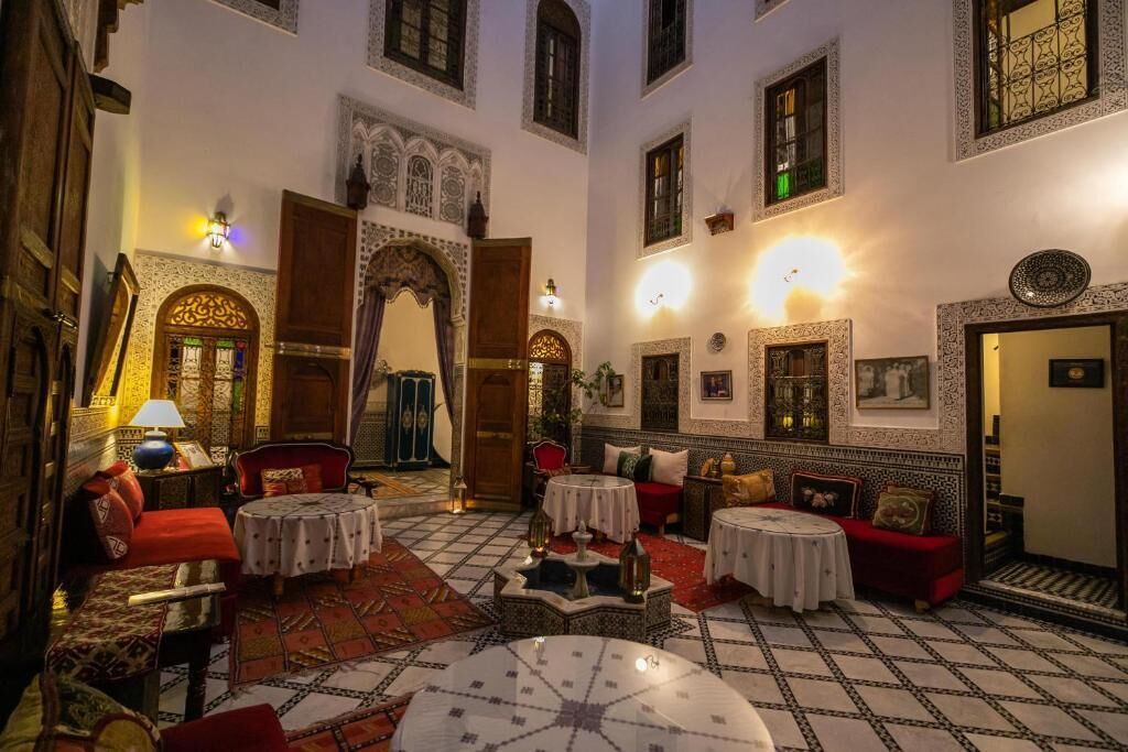 Un interior tradicional marroquí con arcos ornamentados, suelos de azulejos y una variedad de opciones de asiento, incluyendo sofás y mesas.
