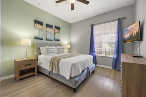 Ein Schlafzimmer mit einem Bett, Nachttischen, einem Deckenventilator, einem Fernseher und einem Fenster mit Vorhängen.