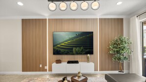 Una sala de estar moderna con un televisor de pantalla plana grande montado en una pared de madera, un mueble blanco para entretenimiento, dos mesitas redondas con jarrones decorativos, una planta grande en maceta y un sofá gris.