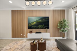 Ein modernes Wohnzimmer mit einem großen Flachbildfernseher an einer Holzwand, einer weißen Unterhaltungsanlage, zwei runden Beistelltischen mit dekorativen Vasen, einer großen Topfpflanze und einem grauen Sofa.