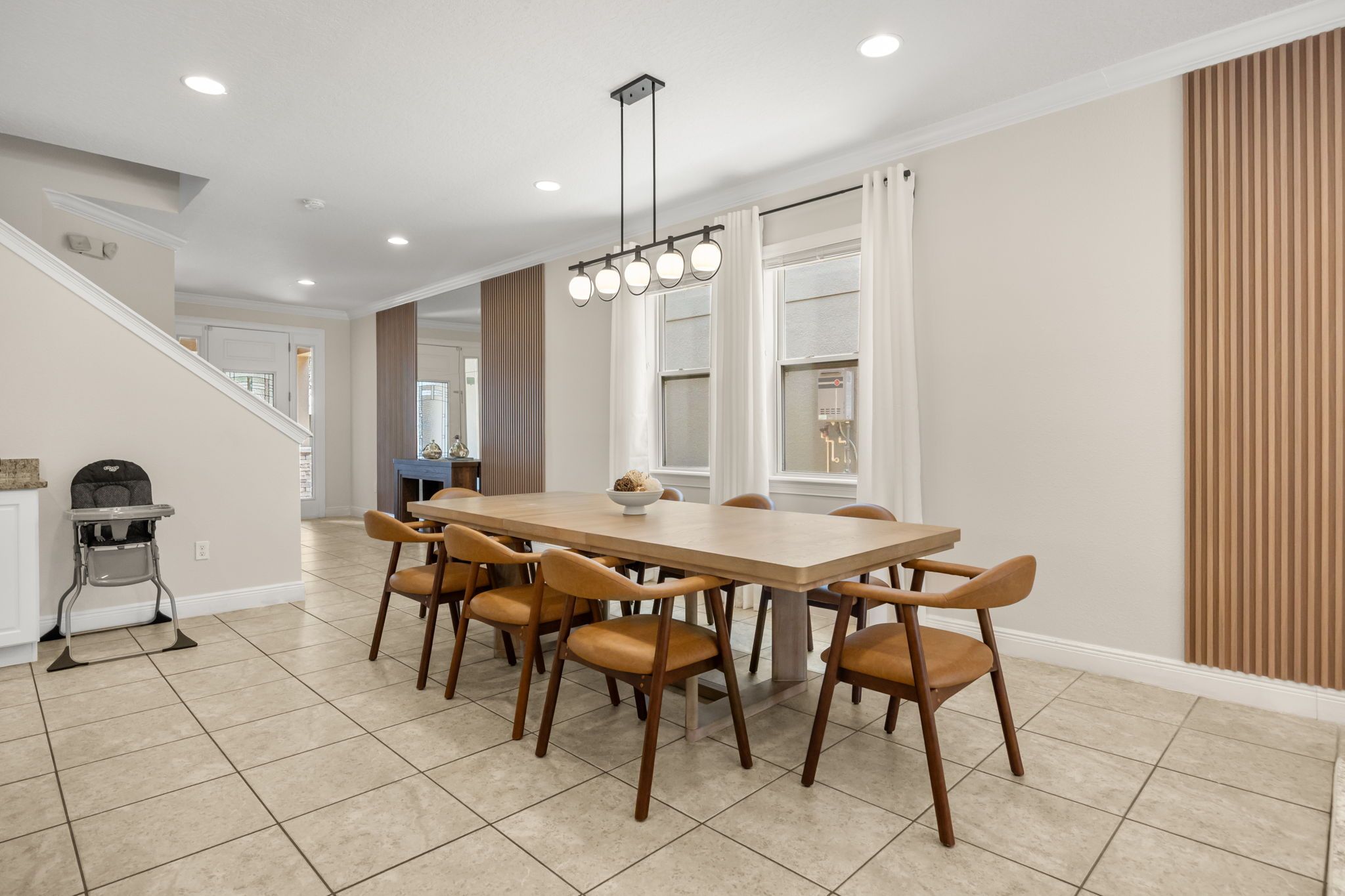 A dining area with a large rectangular table, six chairs, a baby high chair, and a staircase in the background.