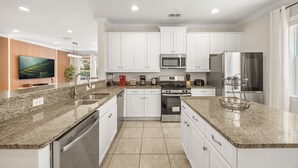 Una cocina moderna con armarios blancos, encimeras de granito, electrodomésticos de acero inoxidable y un televisor de pantalla plana montado en la pared.