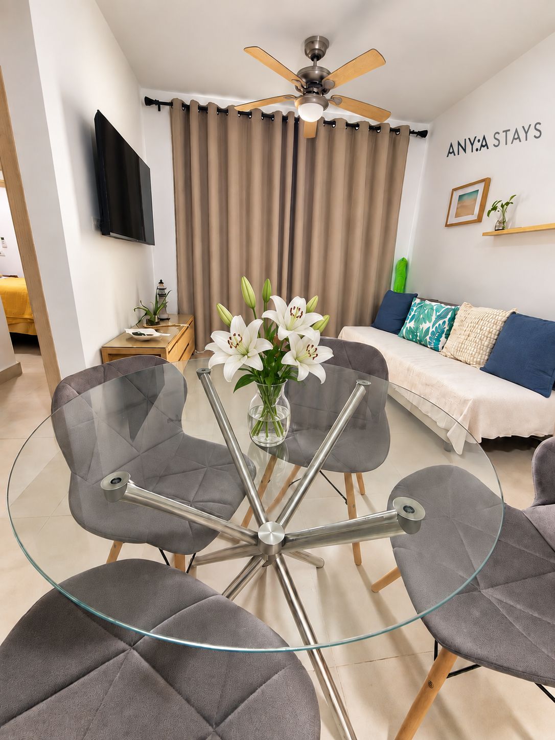 A modern living room with a glass table surrounded by chairs, a ceiling fan, a TV mounted on the wall, and a bed with a blue and white bedspread.