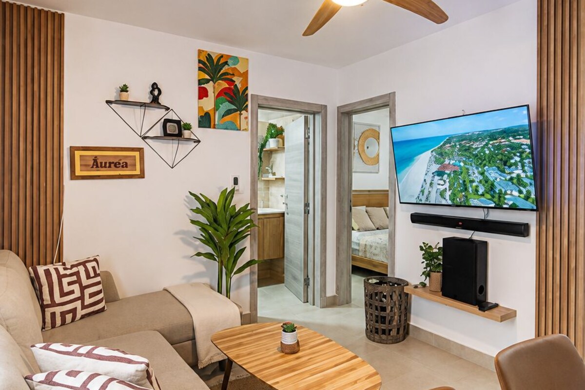 A modern living room with a sofa, coffee table, and chairs, a ceiling fan, a TV, and a plant.