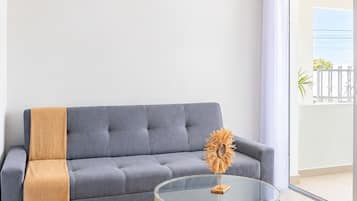 Un salon moderne avec un canapé gris, une table basse en verre avec un socle en bois, un ventilateur de plafond et une grande fenêtre.