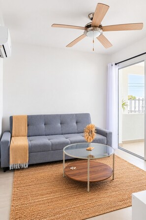 A modern living room with a grey sofa, a glass coffee table with a wooden base, a ceiling fan, and a large window. - ANYA APARTAMENTS (Bayahibe)