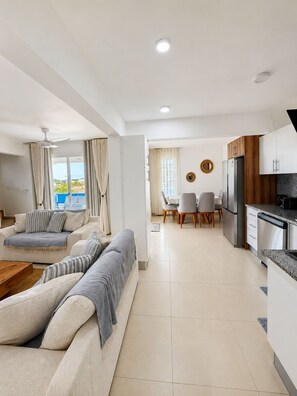 A modern living room with a sofa, a dining area with a table and chairs, and a kitchen with appliances.
