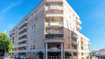 A multi-story hotel with balconies, a central entrance, and a parking area with cars.
