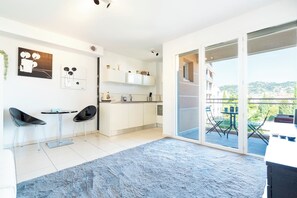 A modern kitchen with white cabinets, a dining area with two black chairs, and a large sliding glass door leading to a balcony with outdoor furniture. - Le Studio du Park avec terrasse et garage (Cannes)