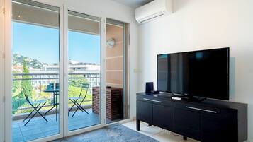 A modern living room with a large flat-screen TV, a sliding glass door leading to a balcony, and a city view.