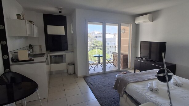 A hotel room with a bed, a desk, a television, a balcony with a table and chairs, and a view of buildings outside. - Le Studio du Park avec terrasse et garage (Cannes)