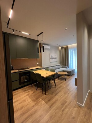 A modern kitchen with a dining area, a grey sofa, and a large window with curtains. - SkyNest by the Lake @30 min to centre, AC and Balcony (Tiranë)