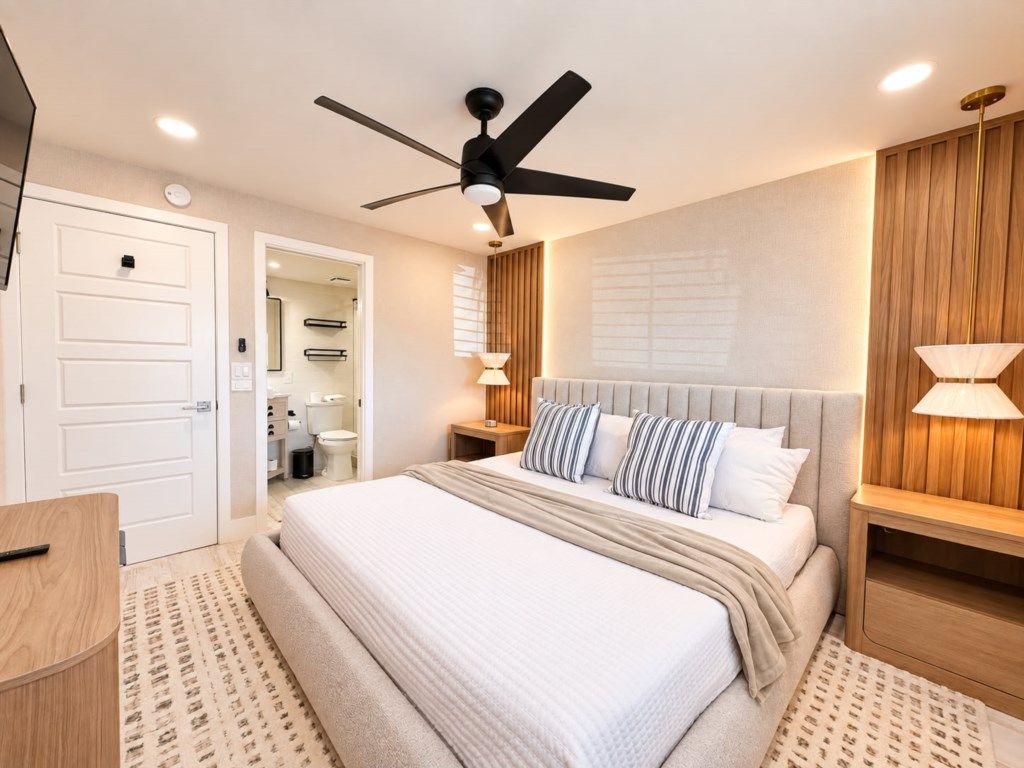 Un dormitorio con cama, ventilador de techo, lámparas de noche, cabecera de madera y una puerta que da al baño.