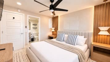 Un dormitorio con cama, ventilador de techo, lámparas de noche, cabecera de madera y una puerta que da al baño.