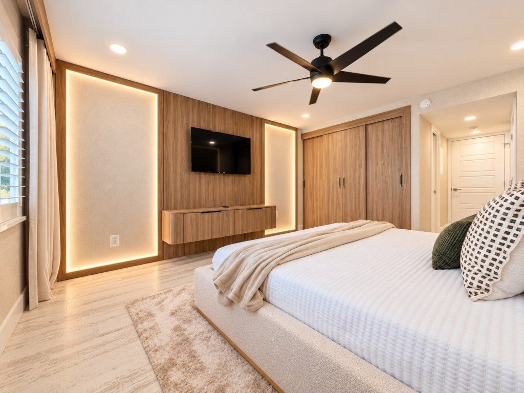 Un dormitorio con cama, ventilador de techo, televisor montado, armarios de madera y una puerta.
