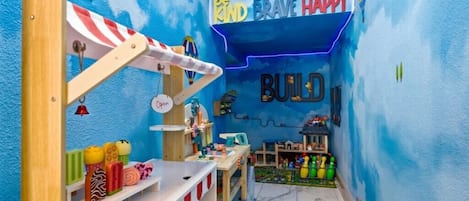 Una sala de juegos con pared azul, una casita de madera y un cartel que dice 'be KIND BRAVE HAPPY'.