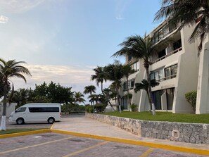 A parking area with a white van, a building with multiple floors and balconies, and palm trees. - Three Bedroom Beachfront Villa (Cancun)