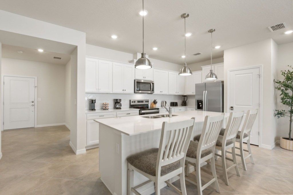 A modern kitchen with white cabinetry, a central island with bar stools, stainless steel appliances, pendant lighting, and a potted plant.