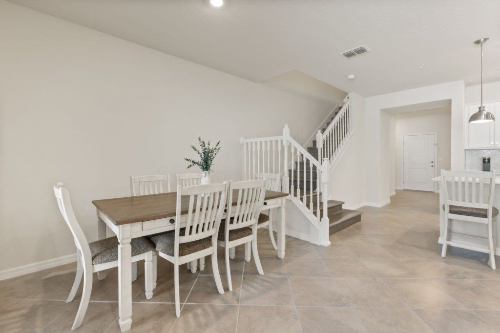 A dining area with a wooden table and white chairs, a staircase, and a kitchen area with white cabinets.