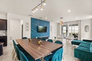 A modern kitchen with a built-in oven, a dining area with a wooden table and teal chairs, and a living area with a sofa and a TV mounted on the wall. - 7 BD / 5.5 BA / Windsor Island Resort 3409 LW (Davenport)