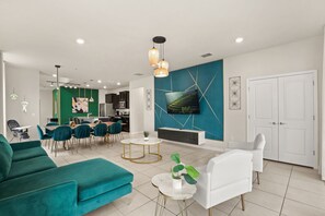 Ein modernes Wohnzimmer mit türkisfarbenen Sofas, ein Essbereich mit blauen Stühlen, eine Küche mit dunklen Schränken und ein großer Fernseher an der Wand.