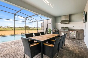 Una cocina exterior con una parrilla, una mesa de comedor con sillas y vista a una piscina.