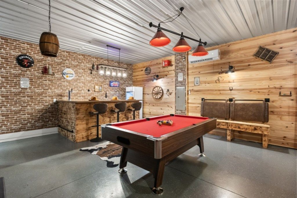 Una mesa de billar con superficie de fieltro rojo, una zona de bar con taburetes y una pared de madera con un reloj y letreros.