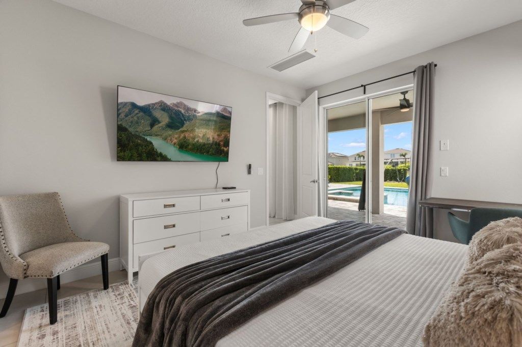 A bedroom with a bed, a chair, a dresser, a ceiling fan, and a large window with a view of a pool and mountains.