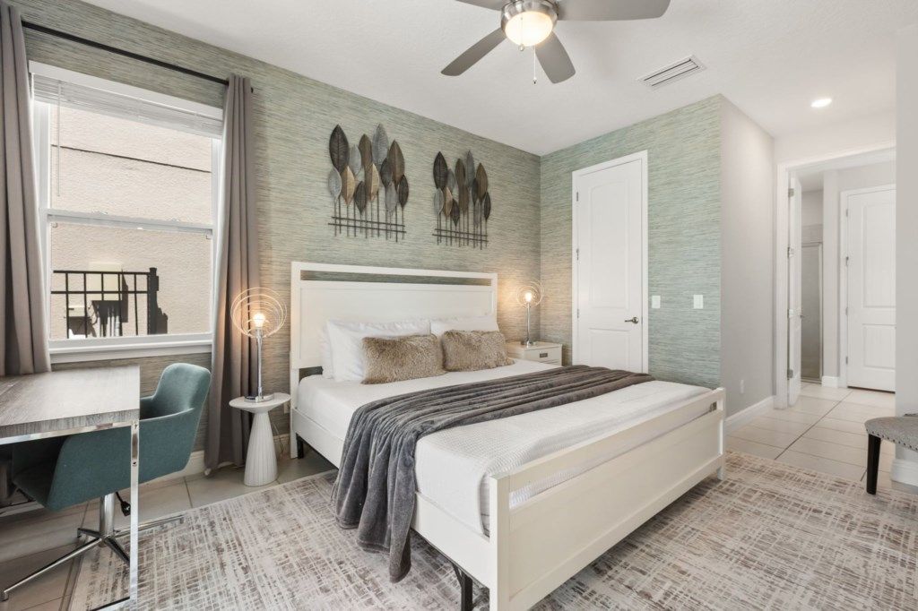 A bedroom with a bed, bedside tables, a desk, a chair, a ceiling fan, and a window with curtains.