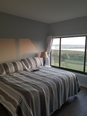 A bedroom with a large bed, a bedside lamp, and a view of a body of water through a window. - Beautiful 2-bedroom condo with AC in Hilton Head Island (Hilton Head Island)