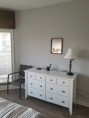 Ein Schlafzimmer mit einem Bett, einer weißen Kommode mit Schubladen, einem Sessel, einer Lampe und einem Spiegel an der Wand.