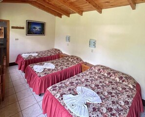Une chambre avec trois lits, chacun recouvert d’une couvre-lit à motifs floraux et doté d’une jupe de lit rouge. Un tableau orne le mur et le plafond est en bois.