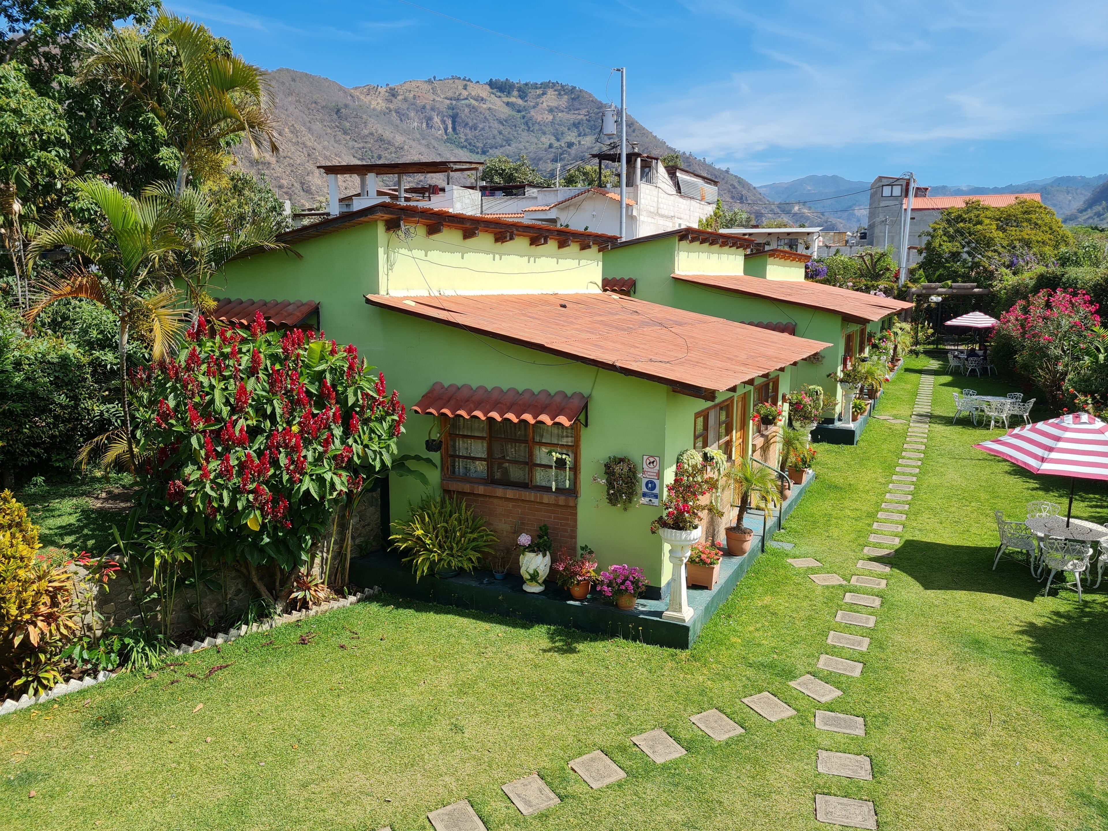 Una casa verde con techo rojo, rodeada de un jardín cuidado y un sendero de piedra que lleva a un área de estar al aire libre.