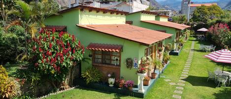 Una casa verde con techo rojo, rodeada de un jardín cuidado y un sendero de piedra que lleva a un área de estar al aire libre.