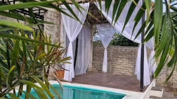 A pool with a white edge, surrounded by a stone wall and a covered seating area with white curtains.