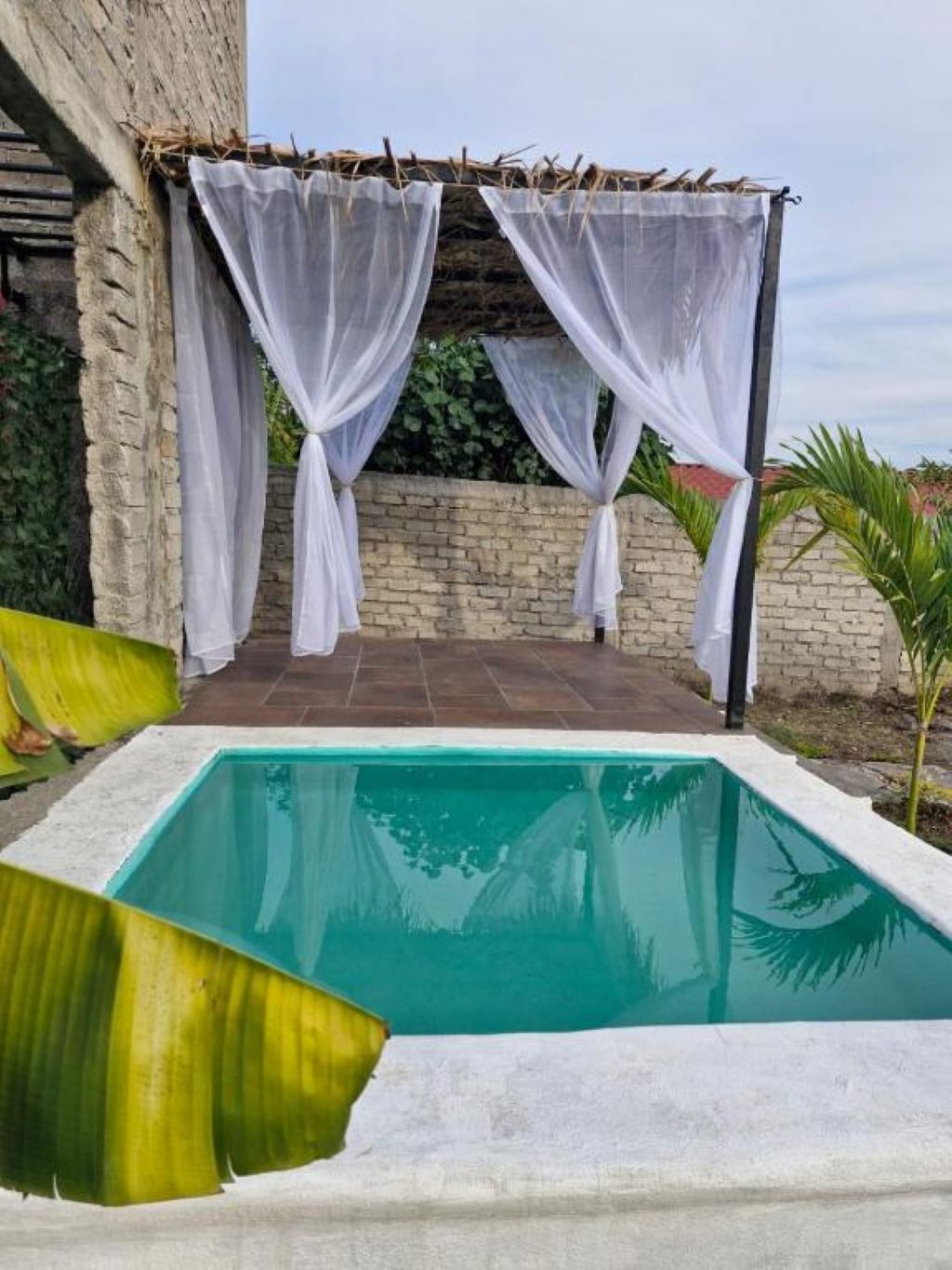 Une piscine agrémentée d’une pergola recouverte d’un tissu blanc, un mur de briques et, au fond, un bâtiment en pierre.