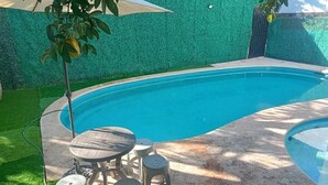 En pool med en böjd kant, omgiven av ett område med grön konstgräs. Där finns ett litet runt bord med fyra pallar och en slang som är ansluten till poolen.