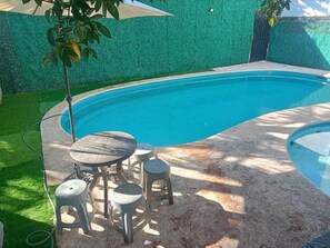 A swimming pool with a curved edge, surrounded by a green artificial grass area. There is a small round table with four stools and a hose connected to the pool. - Depto. con alberca, Aire Acondicionado y cerca de Carrizalillo, hasta 6 personas (Puerto Escondido)