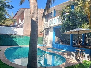 A swimming pool with a curved edge, surrounded by a green lawn and a building with blue and white exterior walls. - Depto. con alberca, Aire Acondicionado y cerca de Carrizalillo, hasta 6 personas (Puerto Escondido)