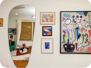 A room with a mirror reflecting a dining area, a window with blinds, and a wall with framed artwork.