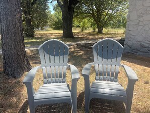 Two white plastic chairs positioned outdoors, surrounded by trees and a stone wall.