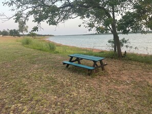 A picnic table in a grassy area near a body of water, with trees and a clear sky in the background. - 3-Acre Wildlife Retreat : Sleeps & Dines 12 : 5 mi to Duncan: 26 mi to Fort Sill (Marlow)