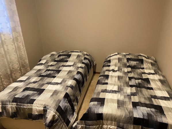Two single beds with a checkered bedspread, a plain wall, and a curtain partially visible.
