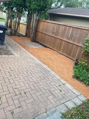 A paved pathway leading to a wooden fence, with a brick patio and mulch on the ground.