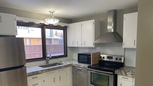 A kitchen with white cabinets, a stainless steel refrigerator, a microwave, a stove, and a window with a view of a wooden fence.