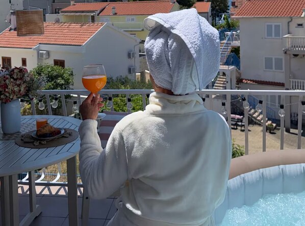 A person holding a glass of orange juice on a balcony with a pool, surrounded by buildings and a clear sky. - One Bedroom Apartment with Sea View and Hydromassage Bath (Vodice)