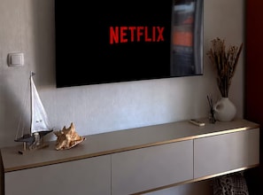 A modern living room with a television displaying the Netflix logo, a small sailboat model, a seashell, and a vase with dried plants on a minimalist console table. - One Bedroom Apartment with Sea View and Hydromassage Bath (Vodice)