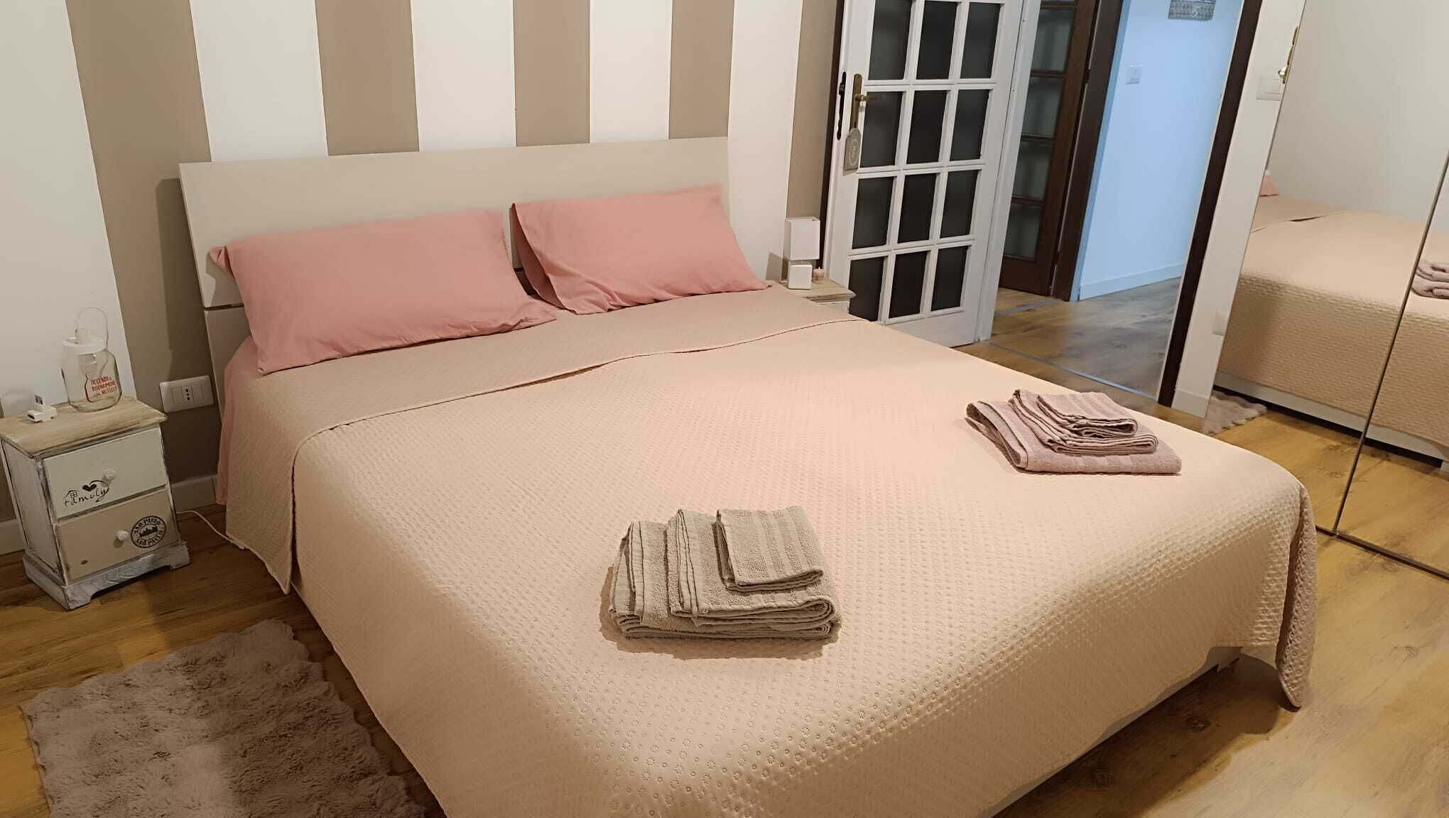 A neatly made bed with pink pillows, a bedside table with a lamp, and a folded towel on the bed.