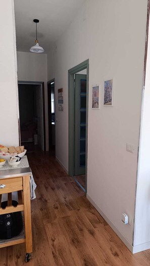 Een gang met houten vloer, een tafel met een broodmand, en een deur naar een andere kamer.